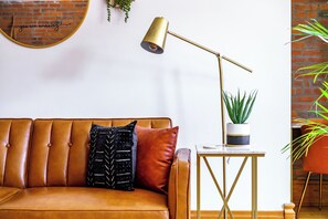A brown leather sofa with a black and white patterned pillow, a gold floor lamp, a small white table with a potted plant, and a brick wall in the background.