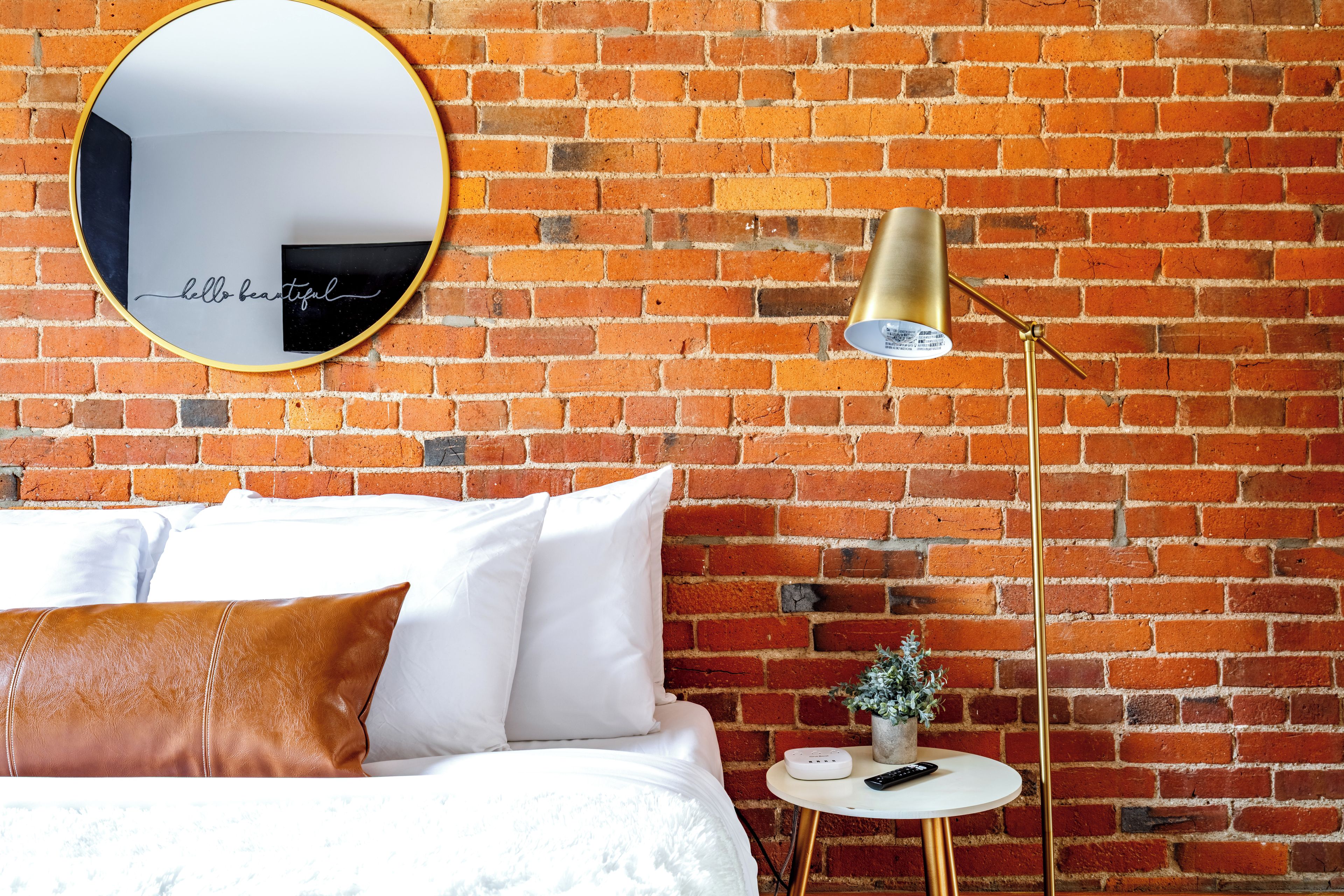 Une pièce avec un mur en briques, un miroir rond, un lit avec une literie blanche, un coussin en cuir marron, une petite table ronde avec une lampe et une plante.
