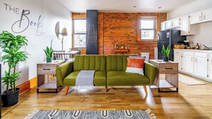 A modern living room with a green sofa, wooden flooring, a brick wall, and a kitchen area with white cabinets. - Jungle House Luxury Lofts – Short North High Street (Columbus)