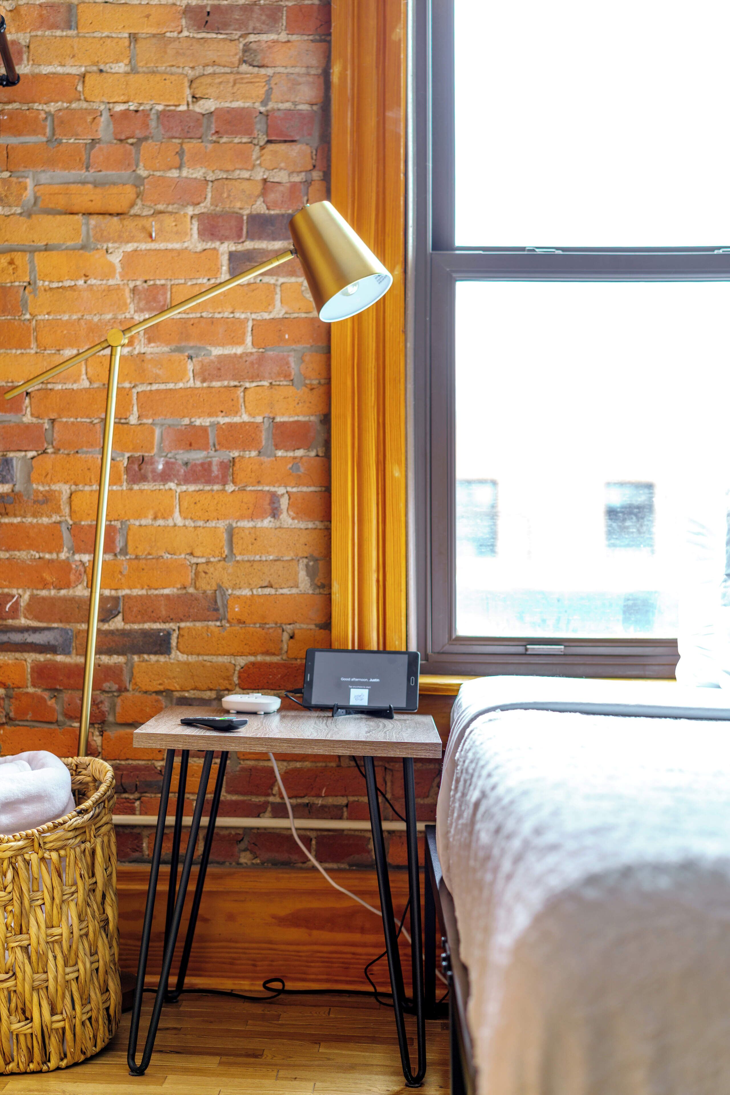 A room with a brick wall, a large window, a bed, a desk with a lamp, and a wicker basket.