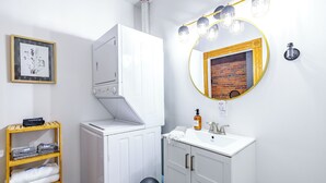 A bathroom with a white cabinet, a wooden shelf with towels, a sink with a mirror, and a wall-mounted light fixture. - Jungle House Luxury Lofts – Short North High Street (Columbus)