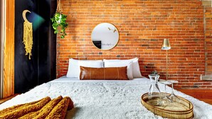 A bedroom with a brick wall, a round mirror, a bed with white and brown bedding, a hanging plant, and a bedside table with a lamp. - Jungle House Luxury Lofts – Short North High Street (Columbus)
