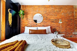 A bedroom with a brick wall, a round mirror, a bed with white and brown bedding, a hanging plant, and a bedside table with a lamp. - Jungle House Luxury Lofts – Short North High Street (Columbus)