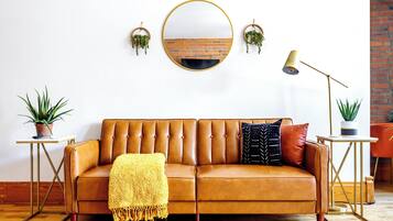 Um sofá de couro marrom com um cobertor amarelo, duas mesinhas de cabeceira com plantas em vasos, um espelho redondo e um abajur.