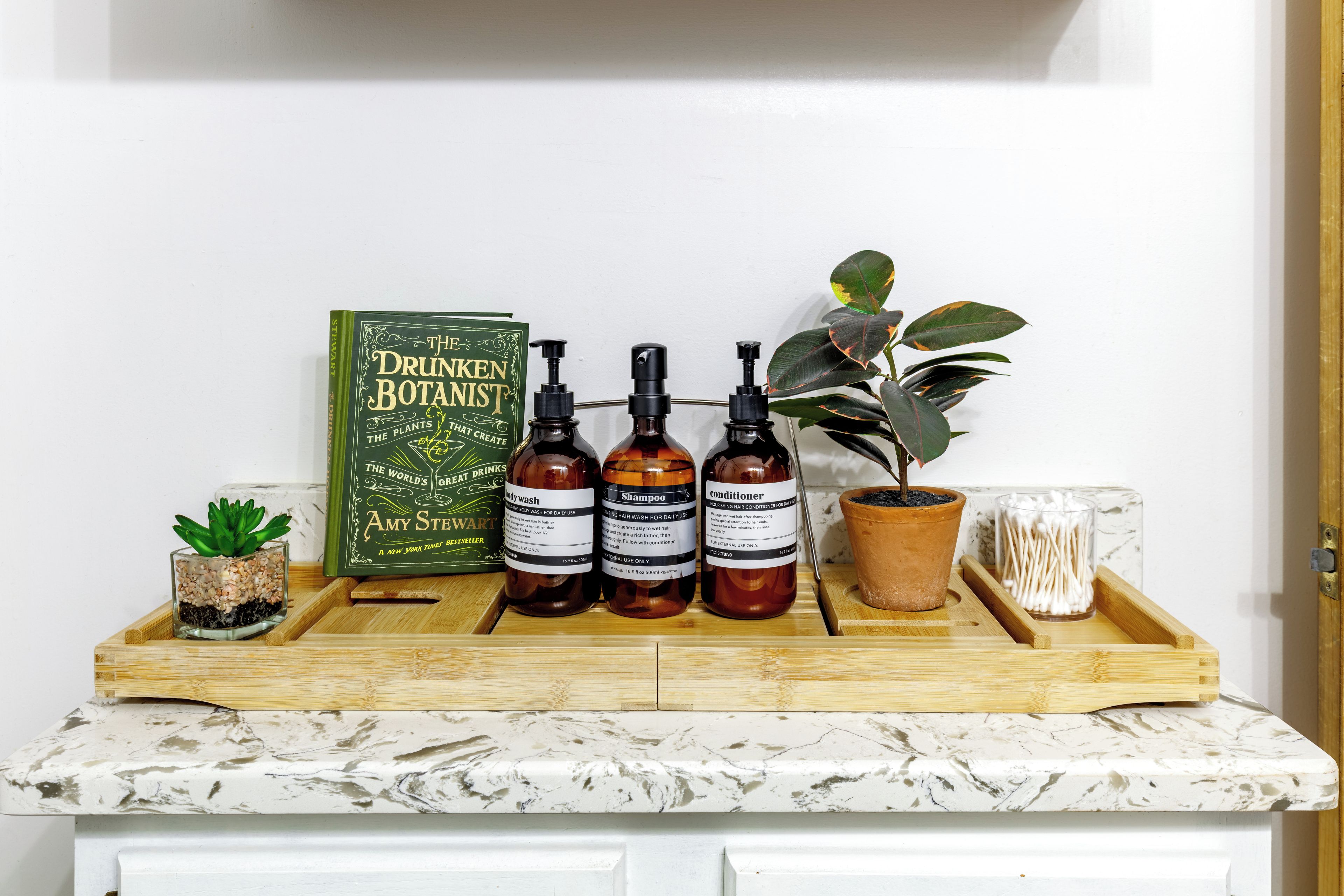 A wooden tray with three bottles labeled 'shampoo,' 'body wash,' and 'conditioner,' a potted plant, a book titled 'The Drunken Botanist,' and a small glass container with cotton swabs.