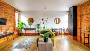 A modern living room with a brick wall, a sofa, a coffee table, a TV, and a dining area. - Jungle House Luxury Lofts – Short North High Street (Columbus)