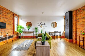 A modern living room with a brick wall, a sofa, a coffee table, a TV, and a dining area. - Jungle House Luxury Lofts – Short North High Street (Columbus)