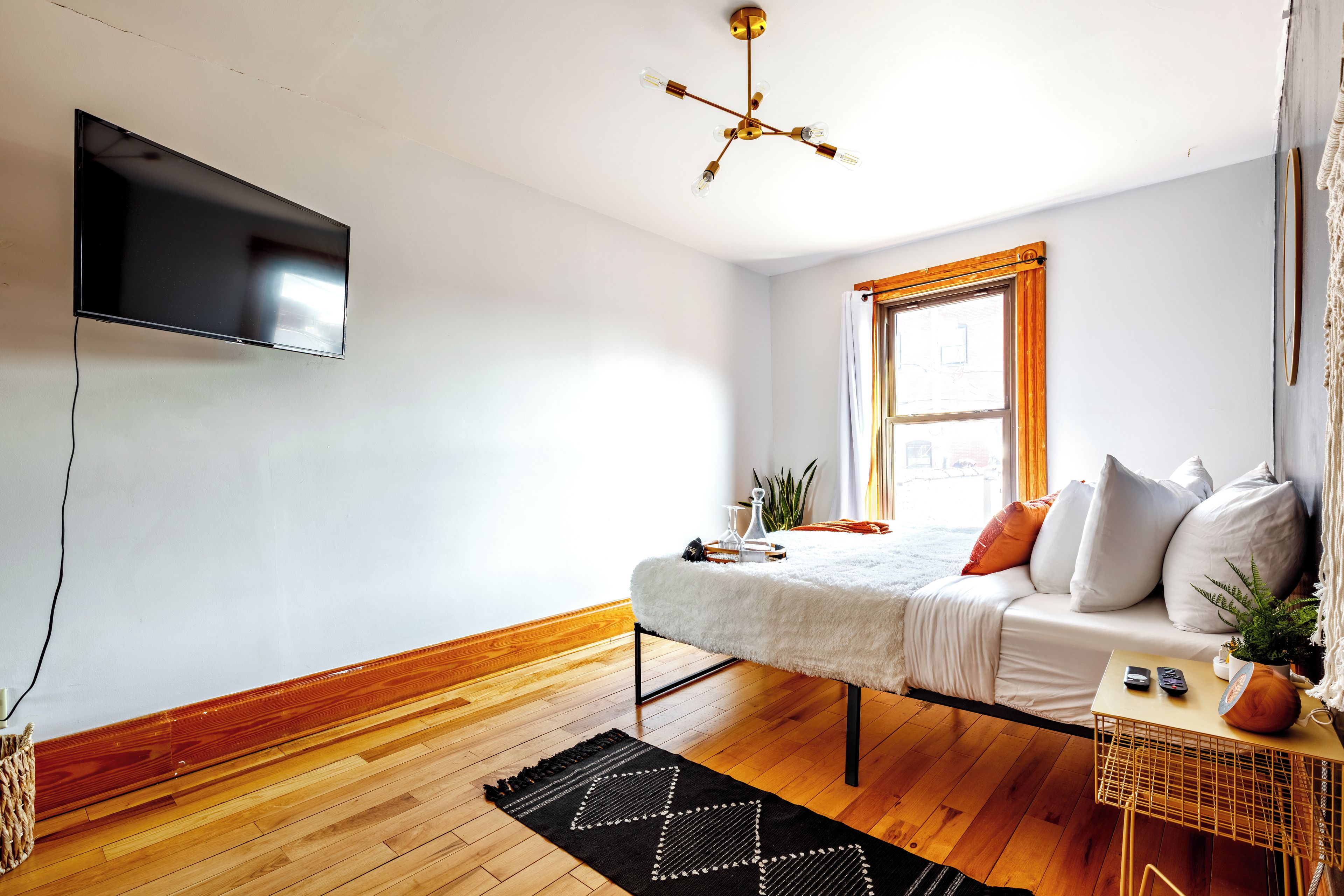 A modern room with a flat-screen TV, a wooden floor, a white sofa with cushions, a small table with a plant, and a window with curtains.