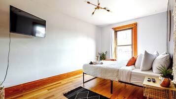 A modern room with a flat-screen TV, a wooden floor, a white sofa with cushions, a small table with a plant, and a window with curtains.