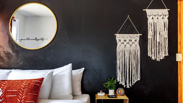 A bedroom with a bed, a round mirror, macramé wall hangings, a small table with a plant, and a wall socket. - Jungle House Luxury Lofts – Short North High Street (Columbus)