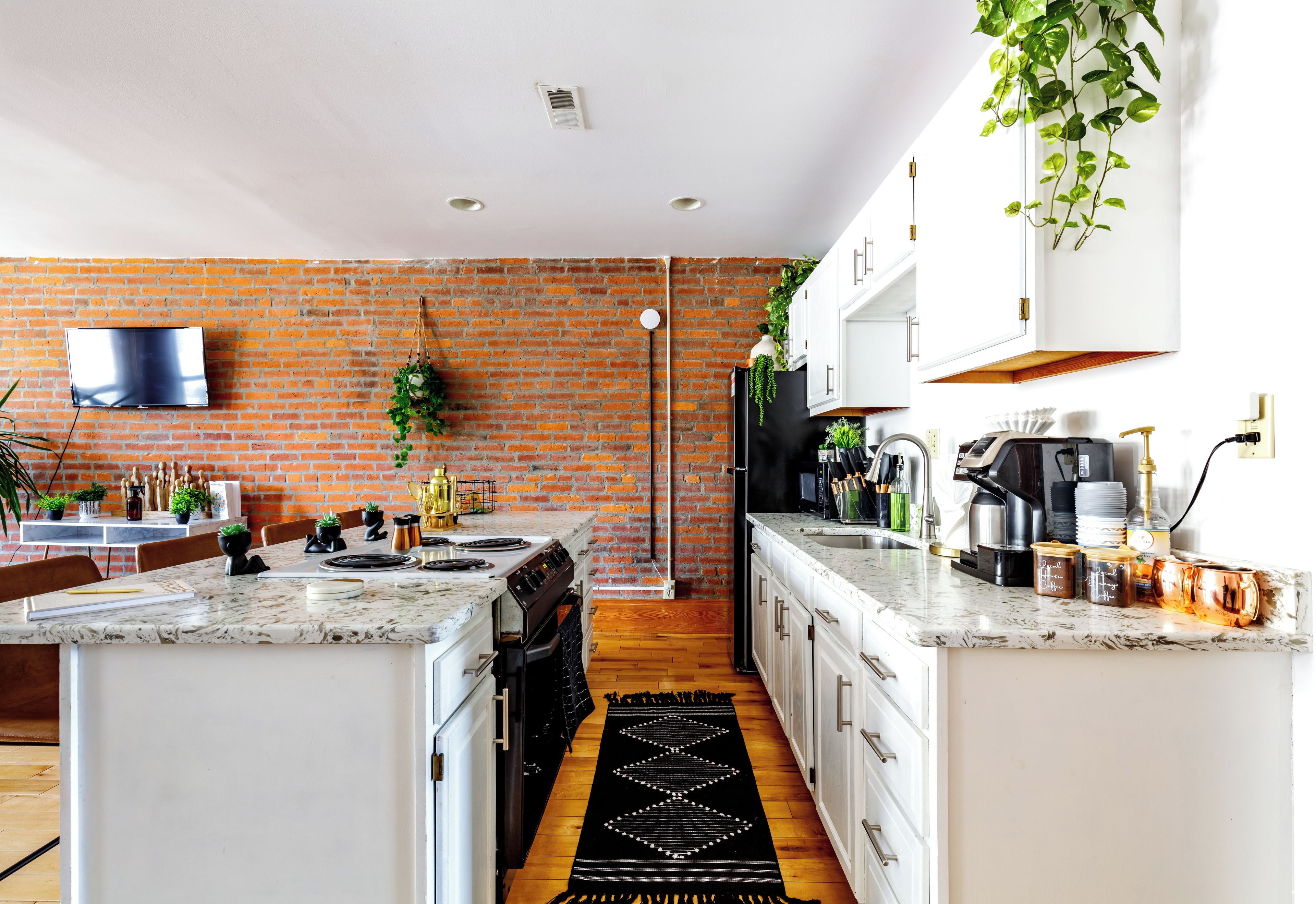 A modern kitchen with a brick wall, white cabinets, a large island, and a flat-screen TV.