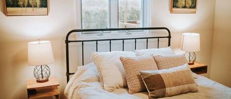 A bedroom with a bed, two bedside lamps, a window, and two paintings on the walls.