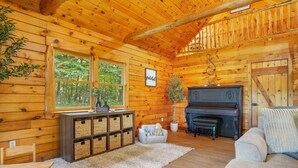 A cozy cabin interior with wooden walls, a piano, a fireplace, and a view of the forest through large windows. - Luxury Retreat in the heart of Perry County, PA. There are plenty of amenities for the whole family to enjoy! (Elliottsburg)
