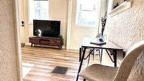 Un loft con piso de madera, televisor sobre un mueble de madera, mesa de comedor con sillas y dos ventanas con persianas.