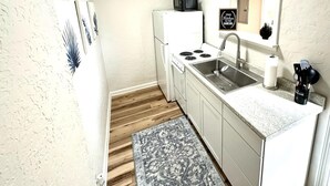 Cocina compacta con gabinetes blancos, estufa, pileta y microondas. Hay una alfombra pequeña en el piso y un cartel en la pared.