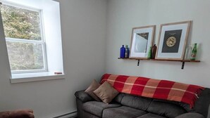 A living room with a grey sofa, a red and white checkered throw blanket, a brown armchair, and a wooden floor. There are two framed pictures on a shelf above the sofa. - The Loft at Spruce Lane (Ingonish)