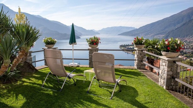A terrace with two chairs, a table, and potted plants overlooking a lake with mountains in the background.