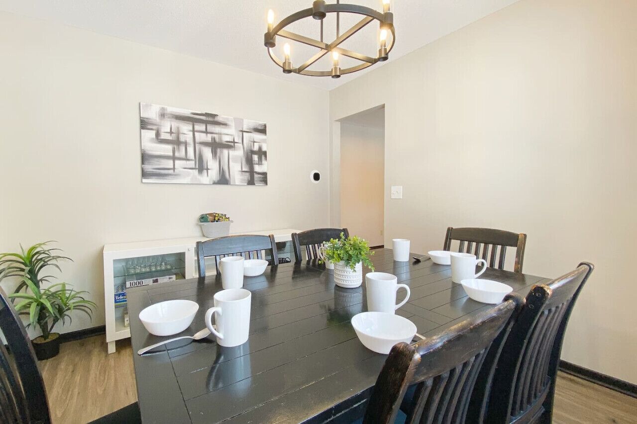 Une salle à manger avec une table en bois foncé, des chaises blanches et un lustre. Un tableau orne le mur et une plante est posée sur la table.
