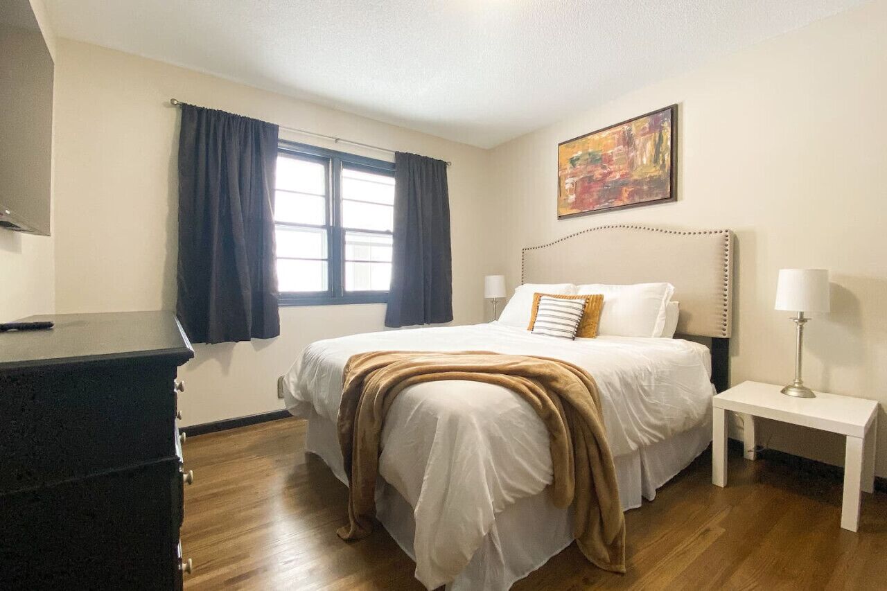 Una camera d'albergo con un letto, comodini, una cassettiera, una finestra con tende e un quadro appeso al muro.
