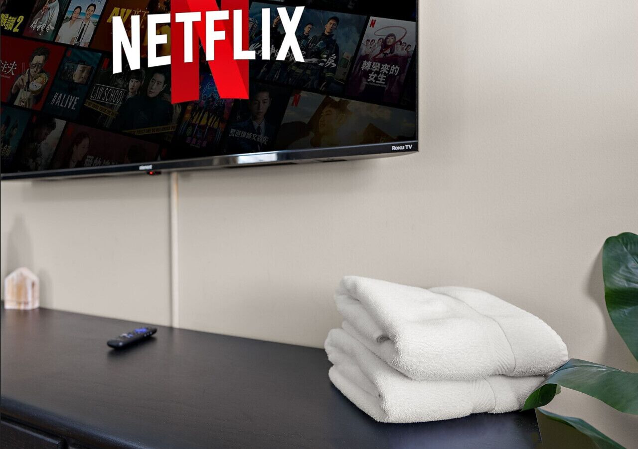 A flat-screen TV mounted on a wall displaying a Netflix interface. A remote control is on a dark surface beside the TV, and a stack of white towels is placed on a table next to a plant.