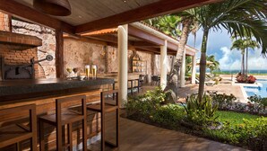 Zona de bar al aire libre con pared de piedra, taburetes de madera y vista a una piscina y palmeras.