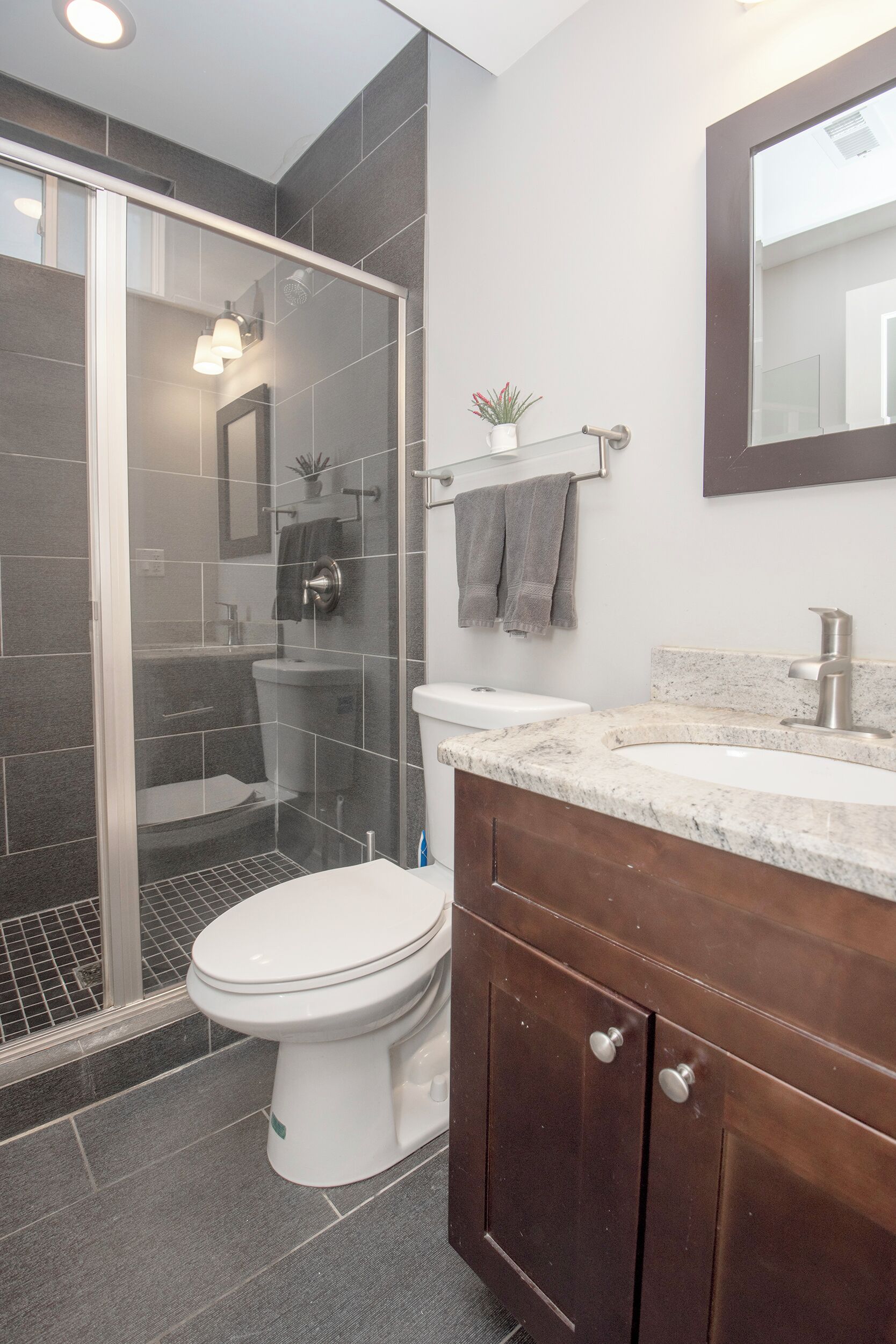 Un baño moderno con ducha envuelta en vidrio, inodoro blanco, lavamanos de mármol y tocador de madera.