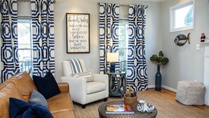 Ein Wohnzimmer mit Sofa, Sessel und Couchtisch. An der Wand hängt ein gerahmtes Schild und es gibt ein Fenster mit Vorhängen.