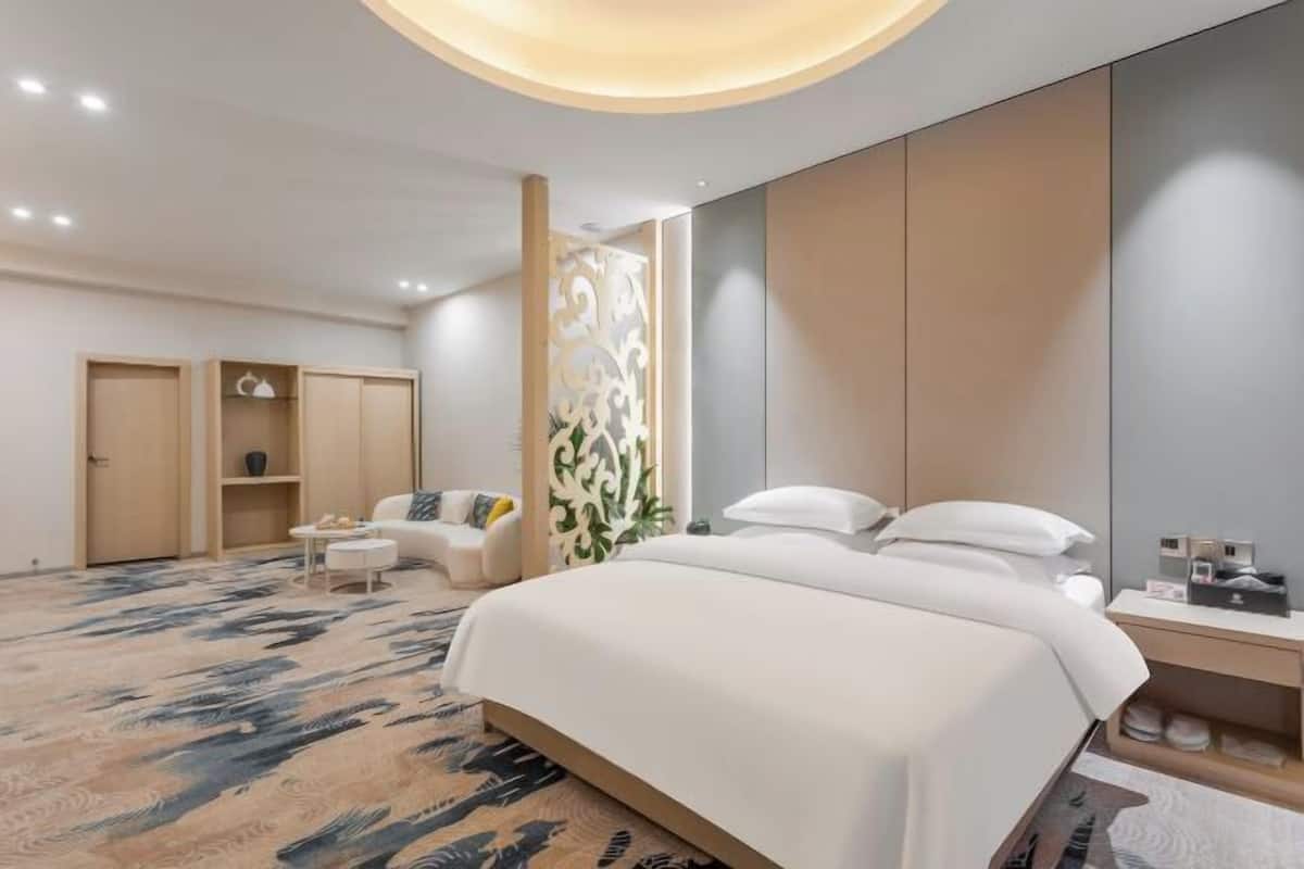 A modern hotel room with a large bed, a seating area with a sofa and coffee table, a wardrobe, and a decorative wall panel.