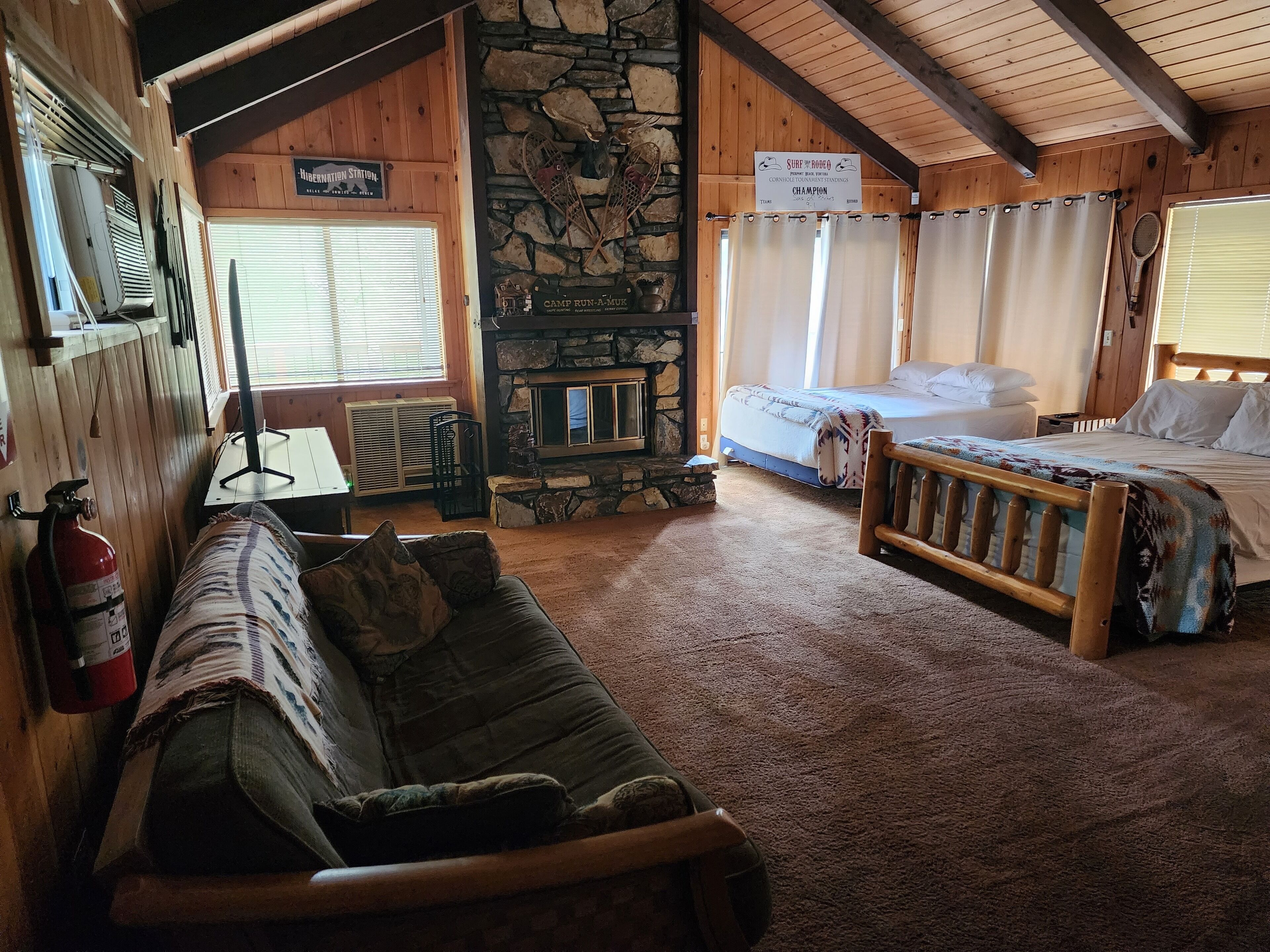Een gezellig kamer in chaletstijl met een stenen openhaard, een bed, een bank en een bord waarop een evenement voor een surf- en touwuitdaging wordt aangekondigd.