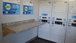 A laundry area with multiple washing machines, a granite countertop, and instructional posters on the wall. - Noble Suites Jacksonville (Jacksonville)