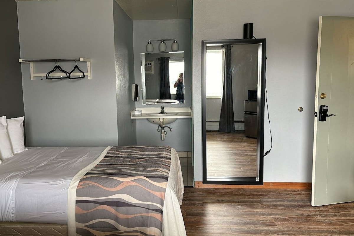 A hotel room with a bed, a bathroom with a sink and mirror, and a view into the kitchen.