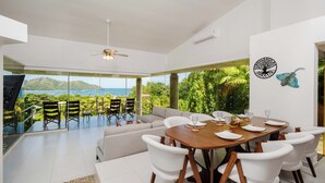 A modern dining area with a wooden table and white chairs, large windows with a view of the sea, and a ceiling fan. - Playa del Coco Hillside Villa • 4BR • Infinity Pool • Bay & Ocean Views (Guanacaste Province)