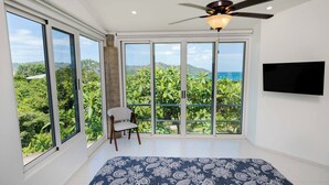 A bedroom with a large bed, a ceiling fan, a TV, and a view of lush greenery and the ocean through large sliding doors. - Playa del Coco Hillside Villa • 4BR • Infinity Pool • Bay & Ocean Views (Guanacaste Province)