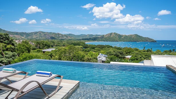 An infinity pool with clear blue water, surrounded by lush greenery and a view of the ocean with boats. - Playa del Coco Hillside Villa • 4BR • Infinity Pool • Bay & Ocean Views (Guanacaste Province)