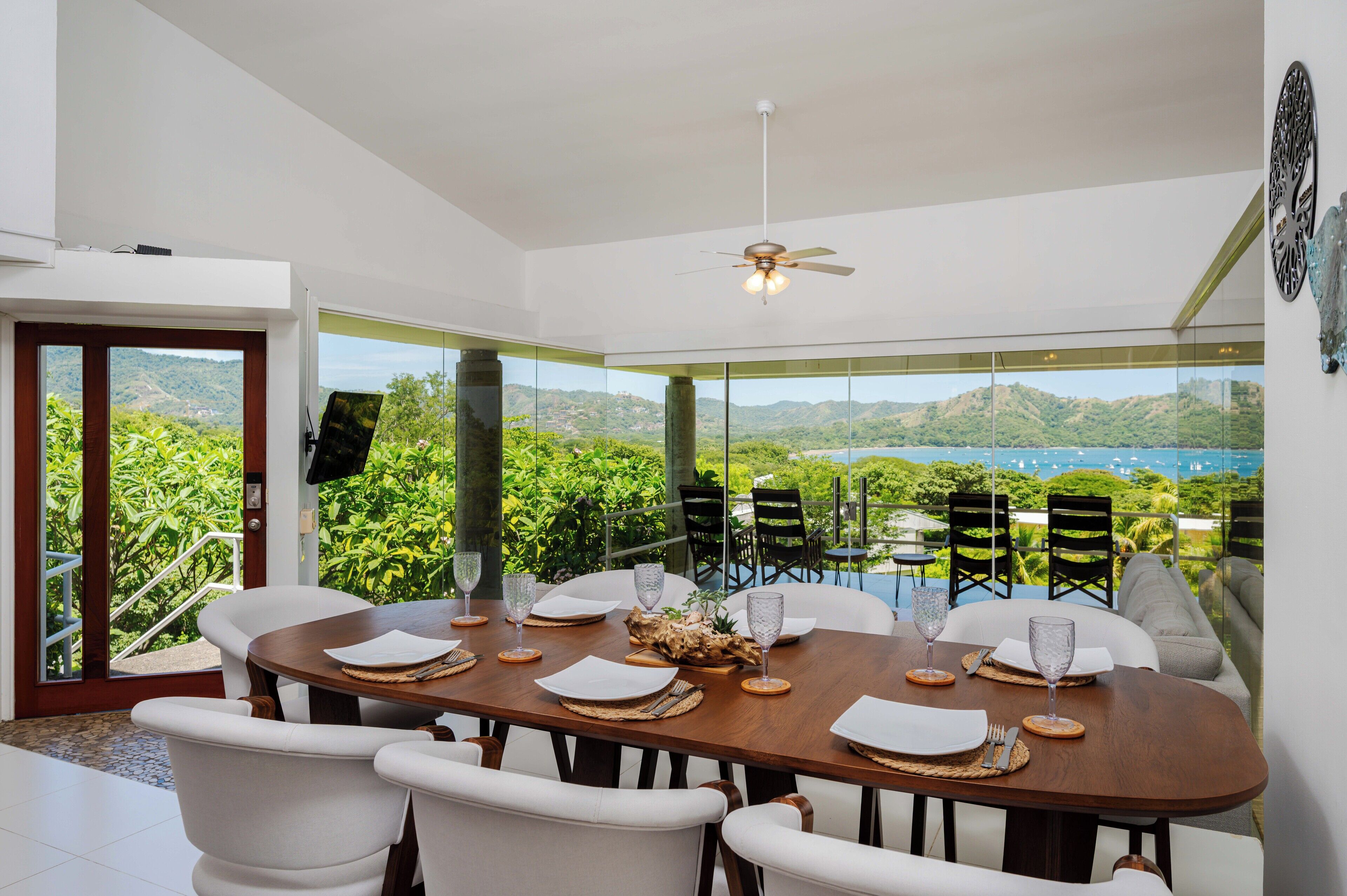 Un coin repas avec une table dressée pour quatre personnes, de grandes fenêtres donnant sur un paysage verdoyant et des collines, et un ventilateur de plafond.