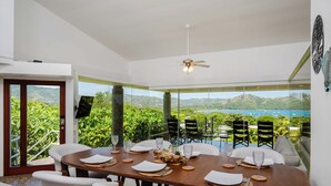 A dining area with a table set for four, large windows offering a view of greenery and hills, and a ceiling fan above. - Playa del Coco Hillside Villa • 4BR • Infinity Pool • Bay & Ocean Views (Guanacaste Province)
