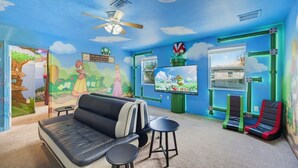 Een kamer met een bank, twee krukken, een plafondventilator, een televisie en een muurschildering met een boom en wolken.