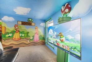 Een kamer met een groot scherm waarop een videospel wordt getoond, een muurschildering met tekenfilmfiguren en een decoratie met Nintendo-thema.