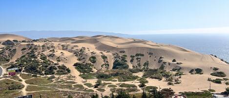 A coastal landscape with rolling sand dunes, a parking area, and a road leading to a beach.