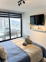 Habitación de hotel con una cama grande, un televisor montado en la pared y vistas a un paisaje urbano a través de amplias ventanas.