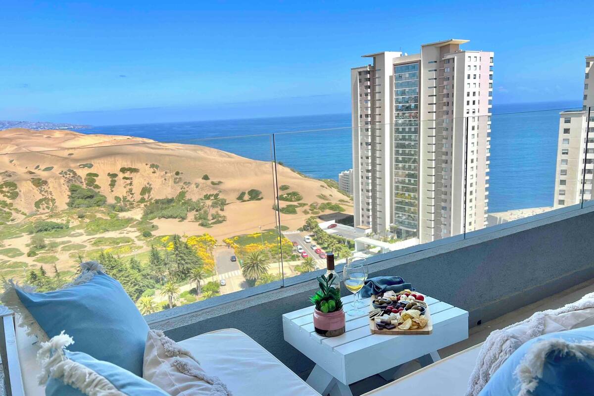 A balcony with a white table set for a meal, a bottle of wine, and a glass. The view includes a sandy landscape and tall buildings.