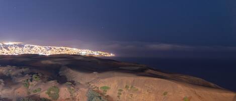 Paisaje nocturno con un horizonte urbano, una colina arenosa y vegetación escasa.