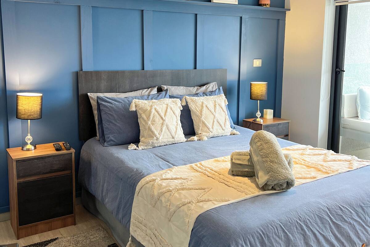 A bedroom with a large bed, two bedside tables, a nightstand, a window with a view, and a ceiling light fixture.