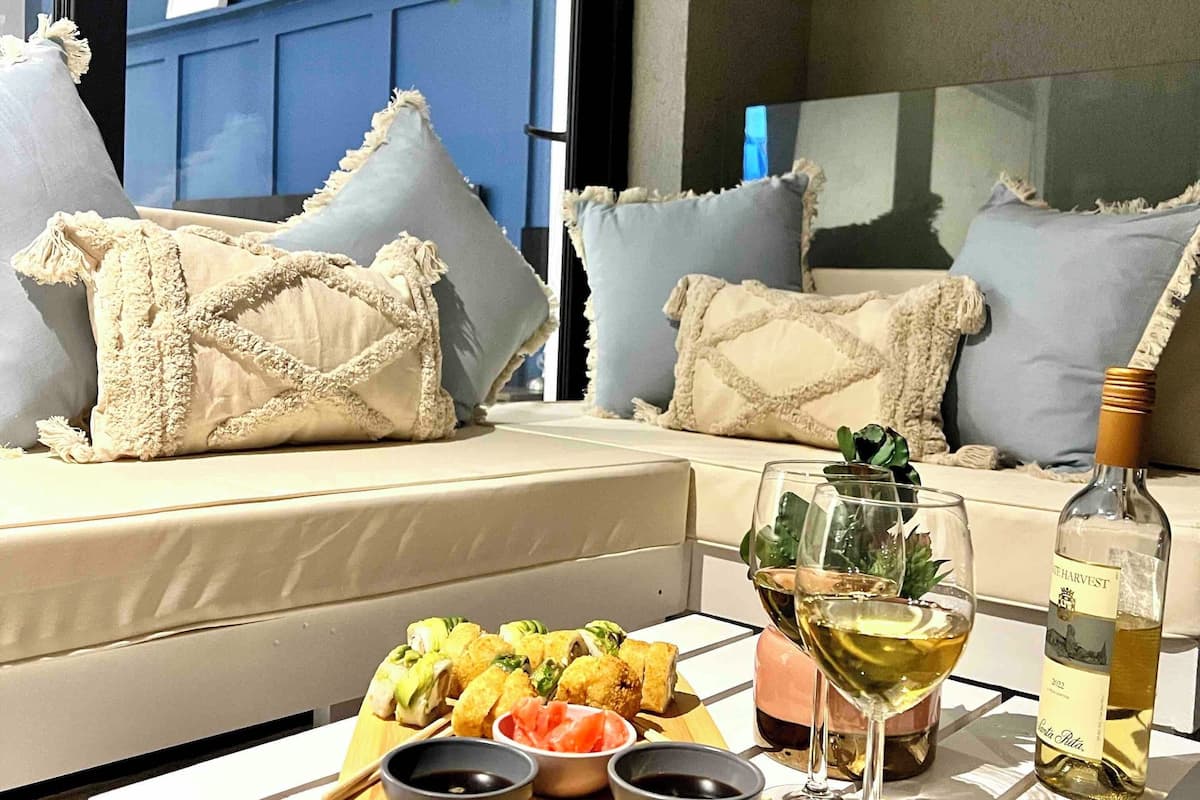 A modern living room with a sofa and cushions, a coffee table with food and drinks, and a view of the outside.