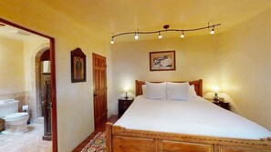 Une chambre à coucher avec un lit en bois, deux lampes de chevet, un tableau encadré au mur et une porte donnant sur une salle de bain.