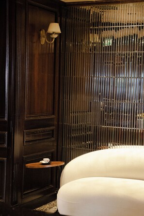A room with a dark wooden panel wall, a white cushioned seating area, a small wooden table with a cup, and a metal-framed window.