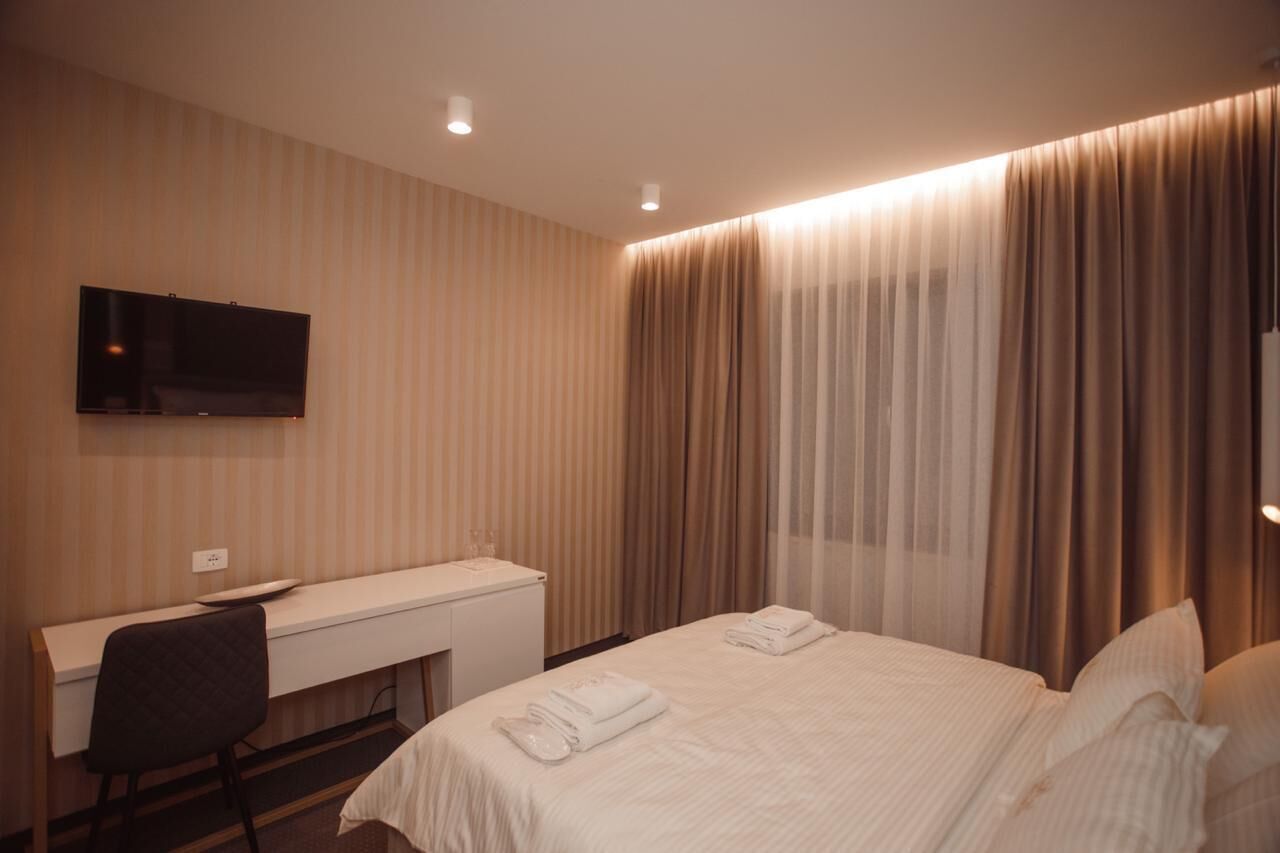 A hotel room with a bed, a desk, a chair, a TV mounted on the wall, and a window with curtains.