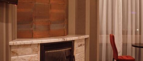 A hotel room with a stone fireplace, a television mounted on the wall, a bed, a chair, and a window with curtains.