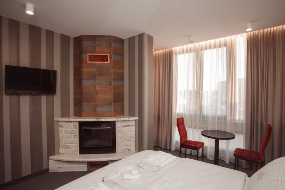 A hotel room with a fireplace, a TV mounted on the wall, a bed with neatly folded towels, a small round table with two red chairs, and large windows with sheer curtains.