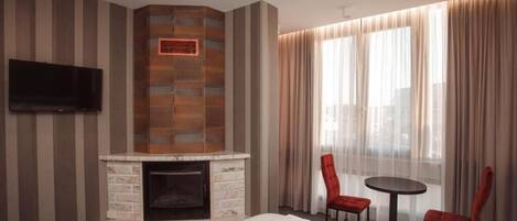 A hotel room with a fireplace, a TV mounted on the wall, a bed with neatly folded towels, a small round table with two red chairs, and large windows with sheer curtains.